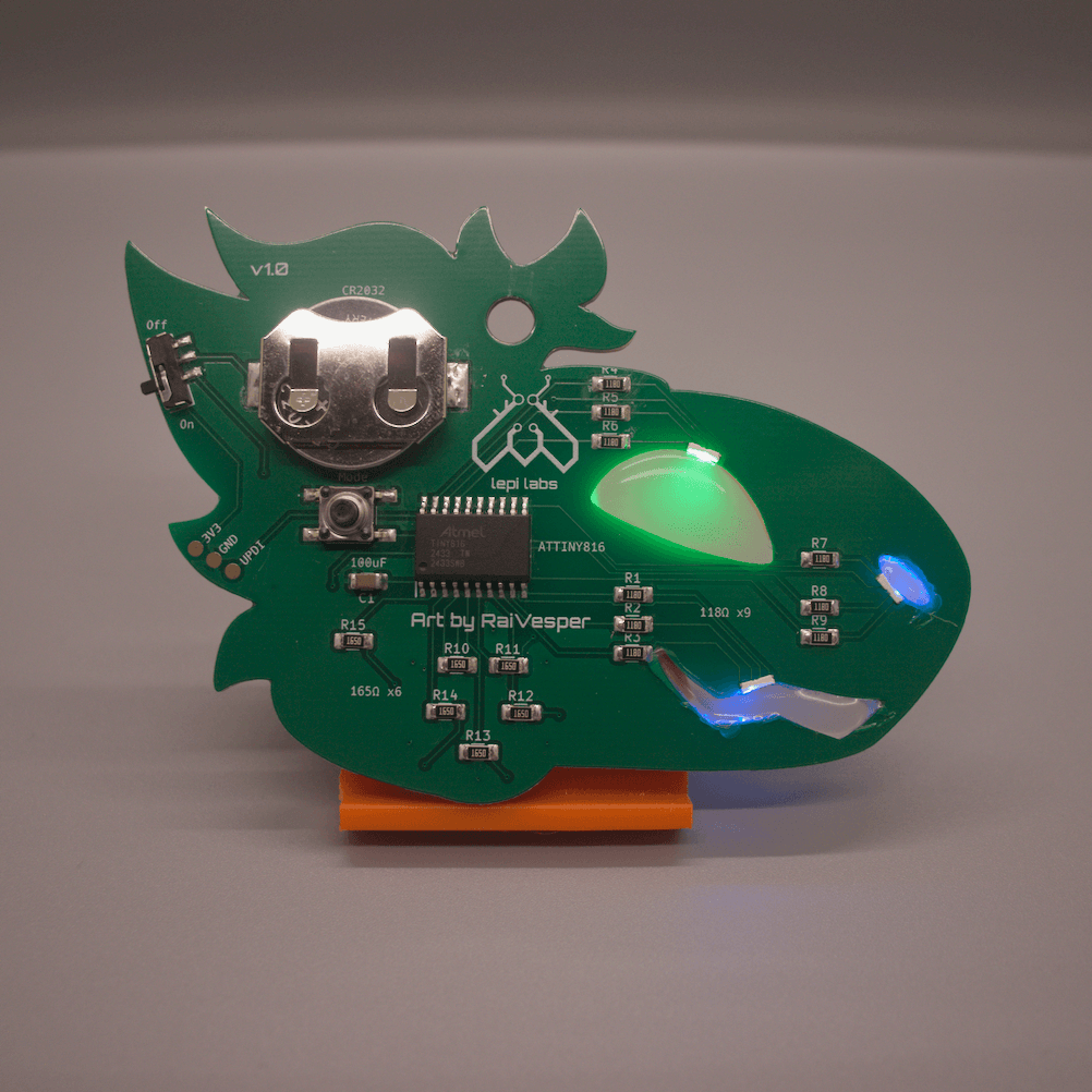 Rear view of a green protogen badge, showing the components which include LEDs, resistors, a coin cell battery, a microcontroller, a push button mode switch and a sliding power switch, and a UPDI connector.